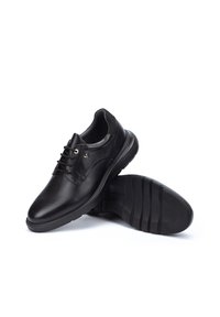 Zapatos de cuero negro con una textura suave, que presentan un cordón en la parte frontal, ojales plateados y una suela de goma con ranuras horizontales.