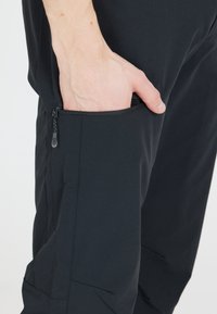 Mustat kangashousut, joissa on vetoketjullinen tasku sivussa. Liukas rakenne, kapea istuvuus ja vahvistettu ommel taskun vieressä.