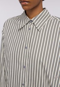 Camicia a righe con strisce verticali bianche e nere, colletto a punta e bottoni blu. Presenta un tessuto morbido e una vestibilità rilassata.