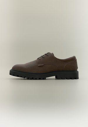Chaussure en daim marron foncé à lacets avec une semelle épaisse en caoutchouc noir crantée, vue de côté sur fond neutre.