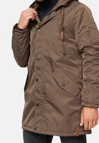 Bruine geïsoleerde parka met een ritszak op de mouw, drukknoopsluitingen en een trekkoordkap. Bevat een gladde, gestructureerde stof.