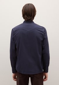 Camisa azul marino de manga larga con cuello puntiagudo, que presenta un diseño de canesú en la parte trasera y puños abotonados. Textura de tela suave.