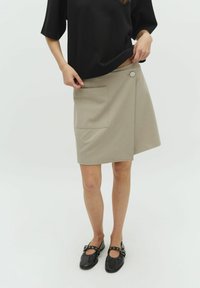 Beige wrap skirt with a side pocket and button closure, made of textured fabric. Worn with a black top and black flats.