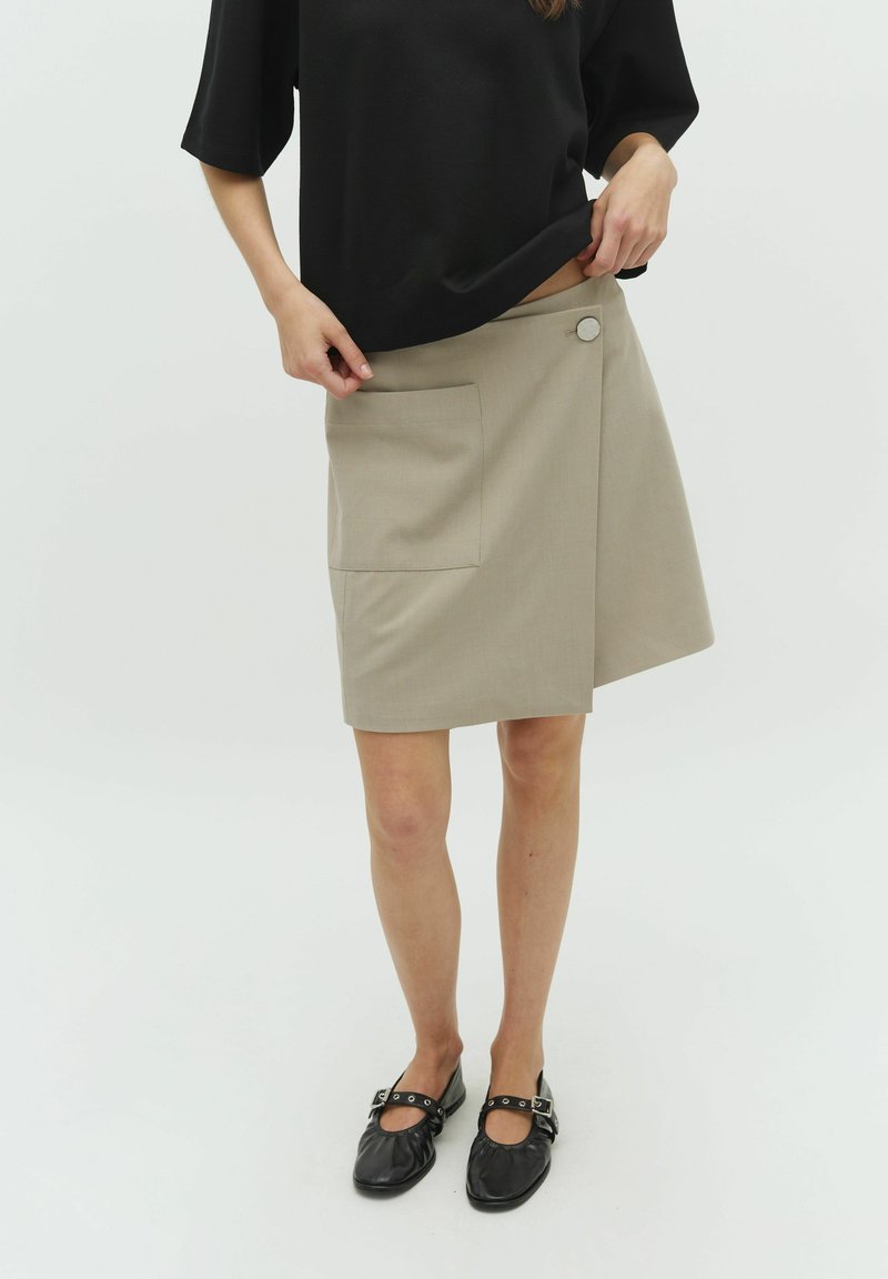Beige wrap skirt with a side pocket and button closure, made of textured fabric. Worn with a black top and black flats.