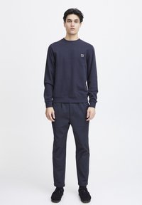 Marineblaue Sweatshirt mit Rundhalsausschnitt und gerippten Bündchen, mit einem kleinen Logopatch. Kombiniert mit marineblauen Hosen und schwarzen Schuhen.