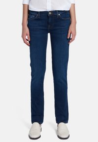 Jean en denim noir délavé avec une coupe slim, légèrement extensible, cinq poches, couture dorée et fermeture à bouton à l'avant, associés à des chaussures blanches.