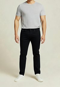 Un homme se tient debout portant un t-shirt gris clair à manches courtes, un jean slim noir et des baskets blanches sur un fond uni.