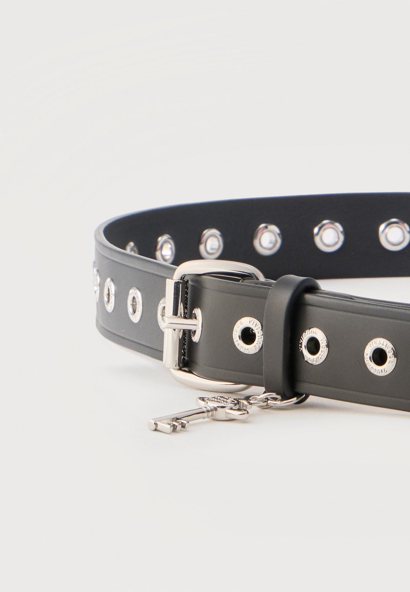 Cintura nera con occhielli e fibbia d'argento, caratterizzata da un piccolo charm a forma di chiave d'argento appeso vicino alla fibbia su uno sfondo semplice.