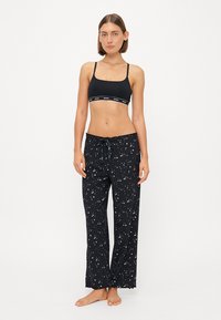 Juodos naktinės kelnės su žvaigždžių raštu ir viršutine dalimi su raišteliu, derinamos su juodu bralette, turinčiu prekės ženklo juostą.