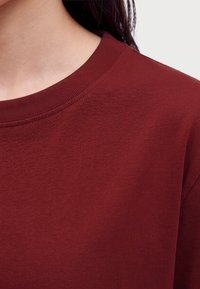 Primo piano di una persona che indossa una maglietta bordeaux tinta unita con scollo rotondo, collo visibile e capelli scuri.