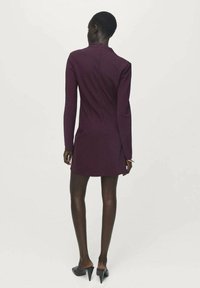 Donna che indossa un vestito mini attillato a maniche lunghe viola scuro e scarpe con tacco nero, in piedi contro uno sfondo bianco e semplice.
