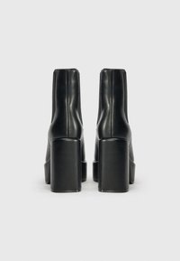 Botines de cuero negro con tacones gruesos altos, mostrados desde la parte posterior sobre un fondo gris neutro.