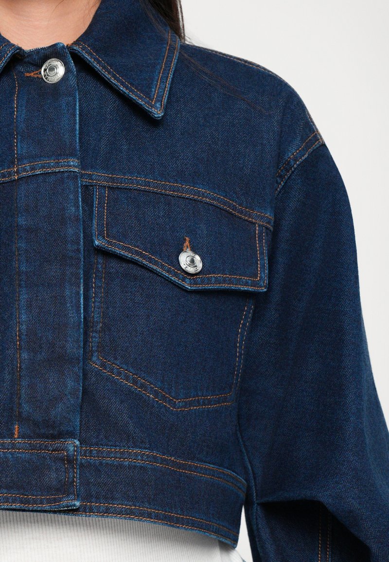 Cropped denim jacket in dark blue with contrasting orange stitching, metal snap closure, and two front pockets. Smooth texture overall.