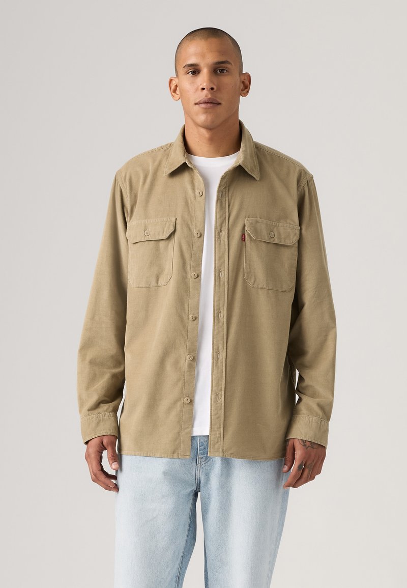 Beige button-down overhemd met twee borstzakken, kraagontwerp en lange mouwen. Draagt over een wit T-shirt, gecombineerd met lichtblauwe jeans.