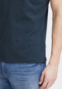 Camiseta de algodón azul oscuro, con cuello redondo, mangas cortas, dobladillo recto y pequeña etiqueta en el costado. Combinada con jeans azul claro.