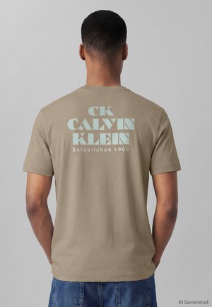 Bărbat purtând un tricou bej cu mâneci scurte cu text albastru deschis "CK Calvin Klein Established 1968" pe spate, asortat cu pantaloni din denim albastru.
