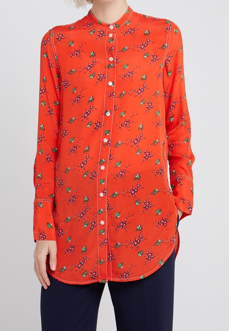 Femme portant un chemisier à manches longues, boutonné, orange vif avec de petits imprimés floraux verts et violets, associé à un pantalon foncé.