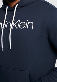 Felpa in cotone blu navy con lacci bianchi e un grande logo frontale "Calvin Klein". Dispone di una tasca a marsupio e un tessuto testurizzato.