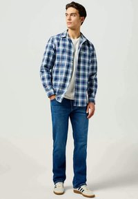 Camisa xadrez azul e branca, usada sobre uma t-shirt branca, combinada com jeans azuis e ténis brancos com riscas pretas.