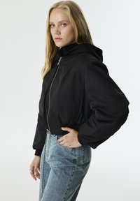 Zwart cropped jack met een capuchon, voorzien van een zilveren ritssluiting aan de voorkant en een elastische tailleband. Gecombineerd met lichtblauwe spijkerbroek.