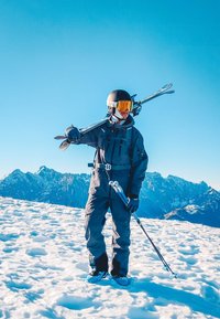 Narciarz w pełnym stroju stoi na śniegu, trzymając narty na ramieniu oraz kijki narciarskie, na tle zaśnieżonych gór pod czystym niebieskim niebem.