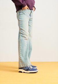 Vaaleansiniset leveälahkeiset farkut, joissa on haalistunut pesu, viiden taskun muotoilu ja näkyvät saumat, yhdistettynä sinisiin tennareihin ja violettiin huppariin.