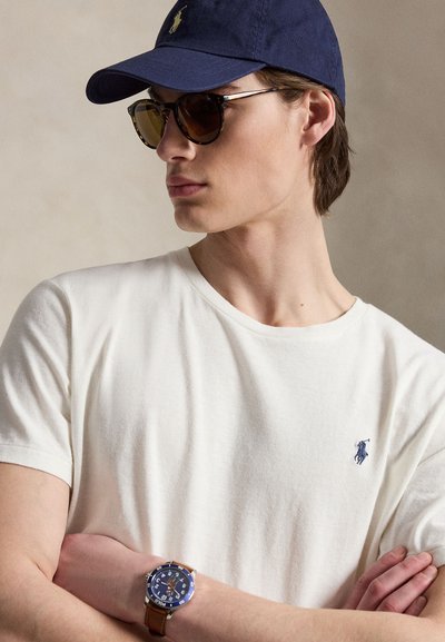 Jonge man met navy pet en schildpadzonnebril, wit poloshirt met logo en horloge met blauwe wijzerplaat en bruine leren band, armen gekruist.