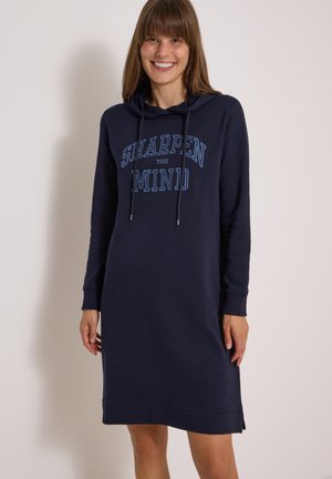 Frau lächelnd, trägt ein marineblaues Kapuzenkleid mit langen Ärmeln und dem Text "SHARPEN YOUR MIND" auf der Vorderseite.