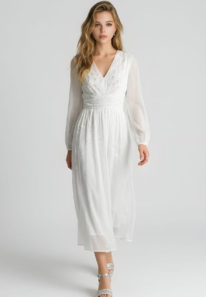 Mujer con un vestido largo blanco con mangas largas transparentes y detalles bordados, combinado con sandalias plateadas de tacón alto, sobre un fondo liso.