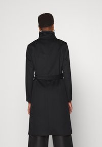 Černý vlněný kabát s vysokým límcem, páskem v oblasti pasu a dlouhými rukávy. Hladká textura a přiléhavý střih, který se vyznačuje minimalistickým designem.