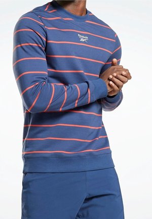 Homme portant un sweat-shirt Reebok bleu avec de fines rayures horizontales rouges, associé à un pantalon bleu, les mains jointes devant lui.