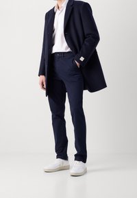 Man i marinblå kappa, vit skjorta, marinblå byxor och vita sneakers står mot en enkel ljus bakgrund.