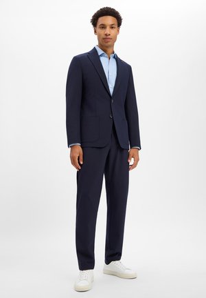 Man standing wearing a navy blue suit with a light blue shirt and white sneakers against a plain white background.