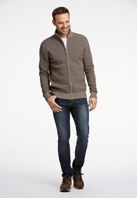 Man med kort hår som bär en brun tröja med dragkedja, blå jeans och bruna sneakers, står mot en vit bakgrund, ler och tittar ner.