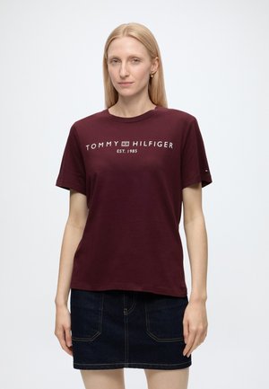 Kvinde med lige blondt hår iført en mørkerød Tommy Hilfiger t-shirt og mørkt denimskørt mod en ensfarvet hvid baggrund.