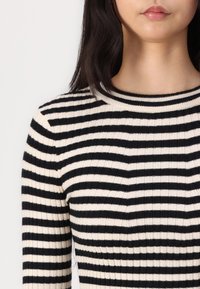 Femme portant un pull côtelé à rayures horizontales noires et blanches, laissant apparaître les épaules, le cou et les lèvres, avec des cheveux raides et foncés.