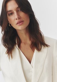 Lichtgekleurde, gestructureerde linnen blazer met een diepe V-hals, voorzien van twee voorknopen en een relaxte pasvorm. Gouden creolen als accessoires.