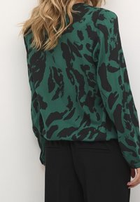 Blouse verte à imprimé animalier noir, manches longues et coupe décontractée. Texture lisse avec un col arrondi et des poignets élastiques. Pantalon noir visible.