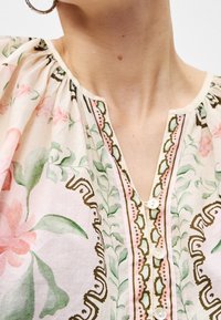 Femme portant un chemisier à motif floral avec des feuilles roses et vertes, col rond avec trois boutons blancs, et une boucle d'oreille en or visible.