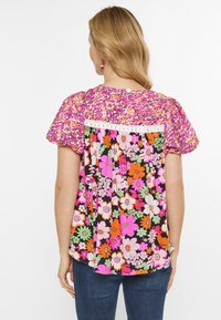 Blomstret top med pufærmer, der har et pink- og sort mønster med orange, grønne og hvide blomster. Trimmet med blonder bagpå.