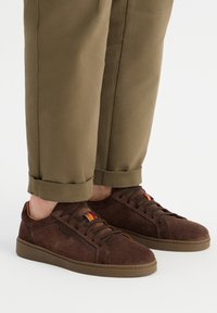 Bruine suède sneakers met een laag ontwerp, vetersluiting en een kleurrijke streepaccent nabij de tong. Rubberen zool, vlak profiel.