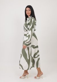 Abito verde e bianco a fantasia con un design fluido, maniche lunghe e vita stretta, abbinato a sandali con tacco trasparente.