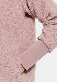 Hellrosa Fleece-Pullover mit strukturierter, flauschiger Außenseite und eng anliegenden Bündchen aus glattem, elastischem Material. Seitliche Taschenanlage sichtbar.