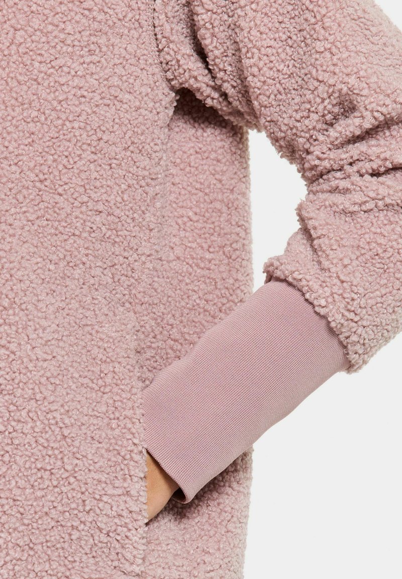 Hellrosa Fleece-Pullover mit strukturierter, flauschiger Außenseite und eng anliegenden Bündchen aus glattem, elastischem Material. Seitliche Taschenanlage sichtbar.