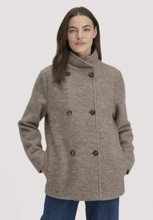 Vrouw in een taupe dubbele stofjas van wol met hoge kraag, met handen in de zakken, staande tegen een effen lichte achtergrond.
