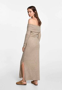 Pletené, off-the-shoulder béžové šaty s dlouhými rukávy a bočním rozparkem. Hladká textura a přiléhavý střih; nošeno s béžovými sandály na klínku.