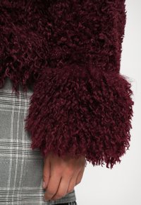 Burgundy fluffy sweater with textured sleeves, featuring loose curls; worn over a gray plaid skirt with a checked pattern.
