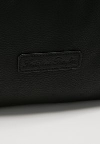 Surface en cuir noir texturé avec un patch noir rectangulaire embossé montrant le nom de la marque cousu "Fritzi aus Preußen."
