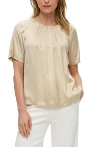 Blouse en soie beige à manches courtes et col froncé, présentant un éclat doux et une coupe décontractée, associée à un pantalon blanc.