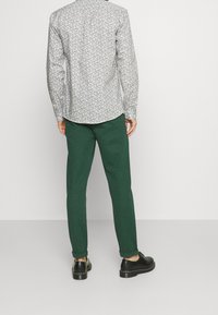 Vit mönstrad långärmad skjorta med rundad nederkant, i kombination med mörkgröna tapered byxor och svarta skor, sedd bakifrån.